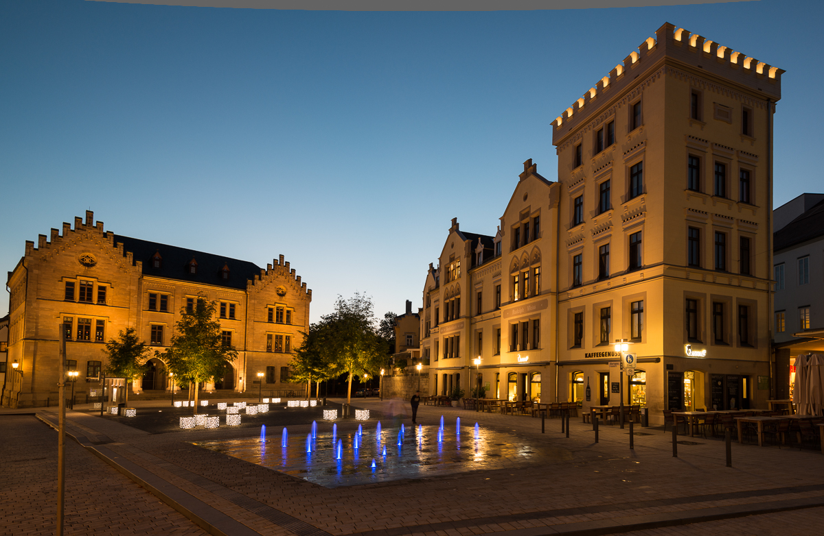Platz mit Wasserspiel am Abend mit bunter Beleuchtung.