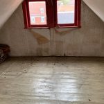 Leerer Raum im Dachgeschoss mit hellen Holzdielen und zwei kleinen Fenstern mit rotem Rahmen unter einer Dachschräge.