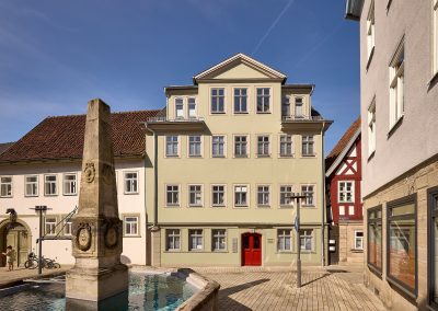 Außenansicht eines hellgrünen Gebäudes mit roter Tür. Im Vordergrund ist ein Brunnen.