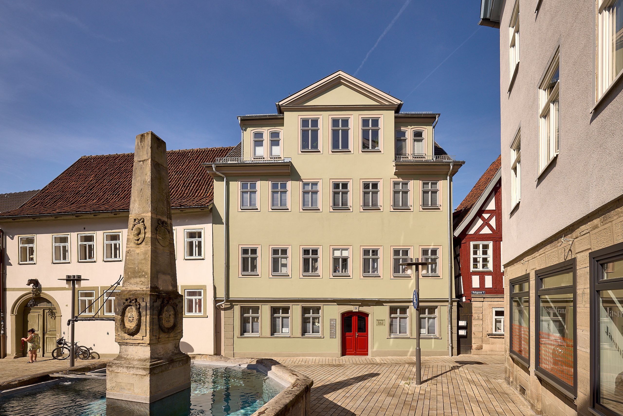 Außenansicht eines hellgrünen Gebäudes mit roter Tür. Im Vordergrund ist ein Brunnen.