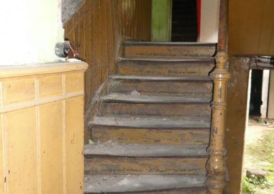 Eine stark abgenutzte, staubige Holztreppe, flankiert von einer gelbbraunen Holzpaneel-Wandverkleidung und einem verzierten Anfangspfosten.
