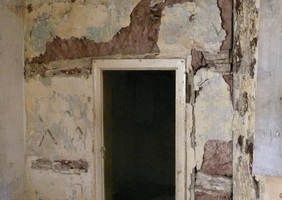 Blick auf einen leeren Türrahmen. Der Putz an den Wänden ist großflächig abgeplatzt und legt das darunterliegende Mauerwerk und alte Holzlatten frei.