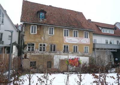 Sanierungsbedürftiges Gebäude mit bröckelnder, gelber Fassade. Ein Banner an der Wand trägt die Aufschrift: „Ich bin ein Denkmal! ...und werde 2016 saniert und restauriert“.
