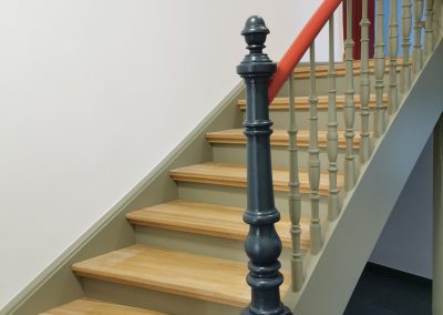 Holztreppe mit grünem Geländer und rotem Handlauf. Der Anfangspfosten des Geländers ist in einem dunklen Grau.