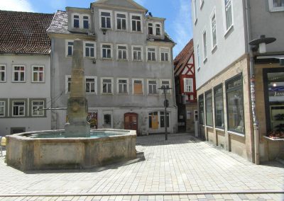 Ein gepflasterter Platz mit einem Steinbrunnen im Vordergrund. Im Hintergrund ein mehrstöckiges, baufälliges, graues Gebäude.